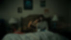 A couple sits on a bed in a dim room. The woman rests her head on the man's shoulder. Beige striped wallpaper and posters in the background.