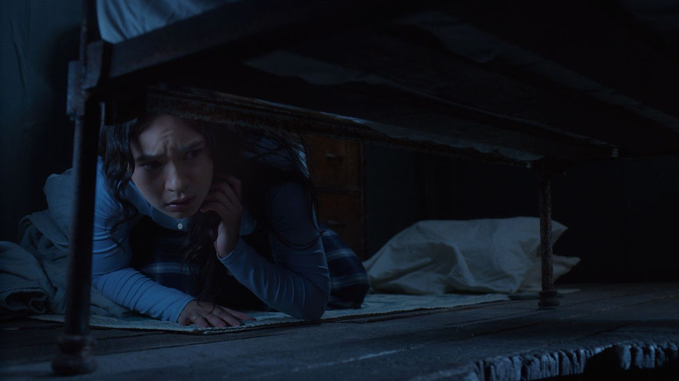 Worried young woman crouches under a bed in a dim blue room, peering out from the shadows.