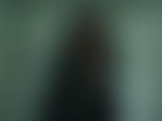 Woman with long dark hair in a black leather jacket stands in a dimly lit room with white brick walls, serious expression.