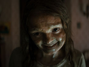 Creepy child with a sly grin in dim room, faintly lit. The background is blurred, creating an eerie and unsettling atmosphere.