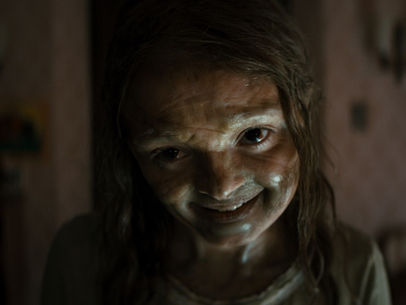 Creepy child with a sly grin in dim room, faintly lit. The background is blurred, creating an eerie and unsettling atmosphere.