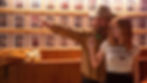 Man in cowboy hat points, guiding woman holding dart. Wall of portraits in the background. She wears 'Horsing Around' shirt. Cozy mood.