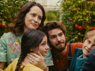 A family of four stands in a lush green garden with tomato plants. They appear joyful and content, the woman wearing a floral shirt smiles gently.