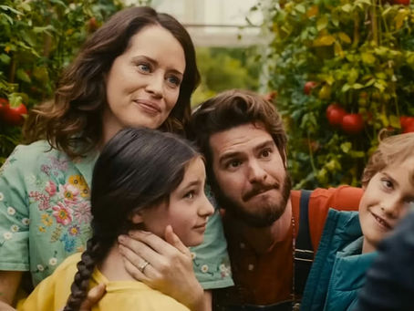 A family of four stands in a lush green garden with tomato plants. They appear joyful and content, the woman wearing a floral shirt smiles gently.