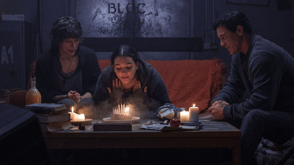 Three people are gathered indoors, one blowing out candles on a cake. Candles, books, and a bottle are on the table. Warm, cozy atmosphere.