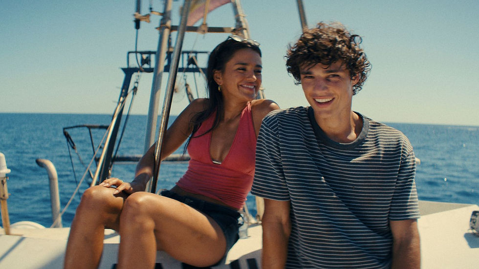 Smiling young couple sitting on a sailboat deck, ocean behind them under a clear blue sky.