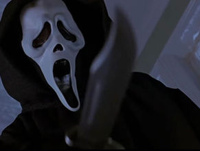 Masked figure in black robe holding knife indoors, against a blue-tinted background. The mask has a white, ghost-like face, evoking fear.