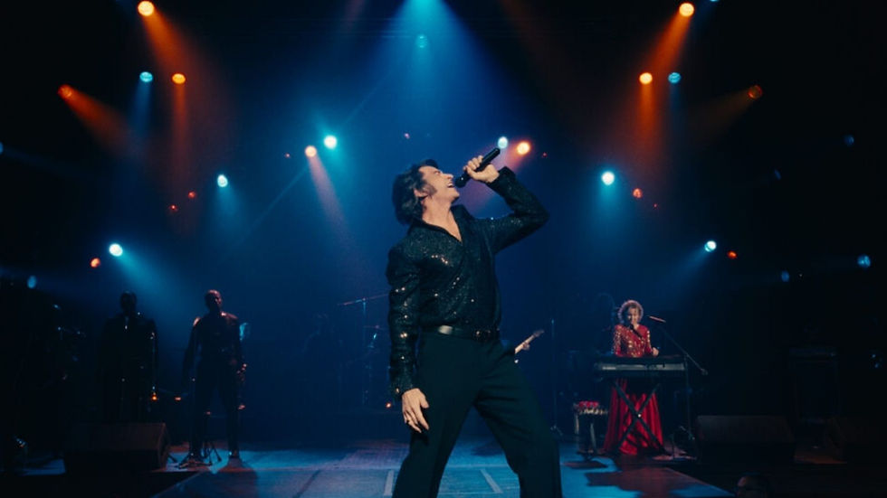 Performer in a glittery shirt sings passionately on stage under colorful spotlights. A keyboardist in red plays on the right. Concert vibe.