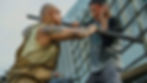 Two men fight on a ship deck, one shoving a metal bar into the other’s chest; both grimace amid railings.