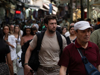 A crowded street scene with people walking. A man in a beige shirt carries a backpack, looking focused. Shops line the background.