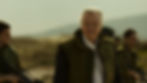 An older man in a green vest stands outdoors with armed guards nearby. The background shows blurred hills under a cloudy sky, creating a tense mood.
