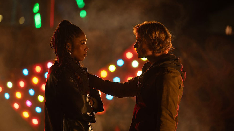 Clayface (2026): Thomas Rhys Harries and Naomi Ackie face each other at night, one touching the other's shoulder, with colourful blurred lights glowing behind them.