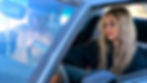 Two people in a car, driver shirtless with a focused expression, passenger with wavy blonde hair looking outside. Bright daylight.