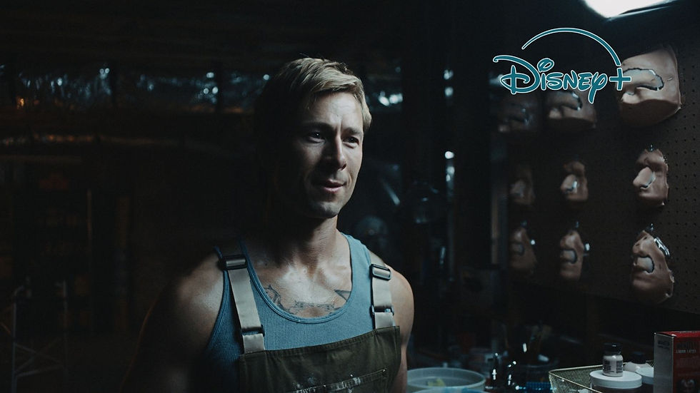 Man in a dim workshop wearing overalls, with masks on the wall and Disney+ logo, looking serious.