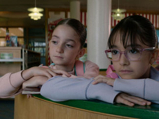 Two young girls, one with pink glasses, rest their heads on a library counter, looking thoughtful. Bookshelves and soft lighting in the background.