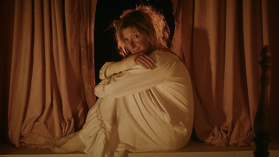 Old woman sitting in white gown with arms crossed staring forward. Sat in front of pink curtains.