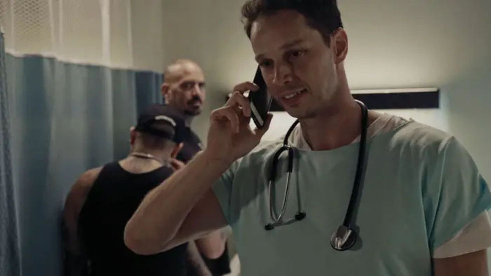 A man with a stethoscope talks on the phone in a hospital hallway. Two men in dark clothing are in the background, one looking back.