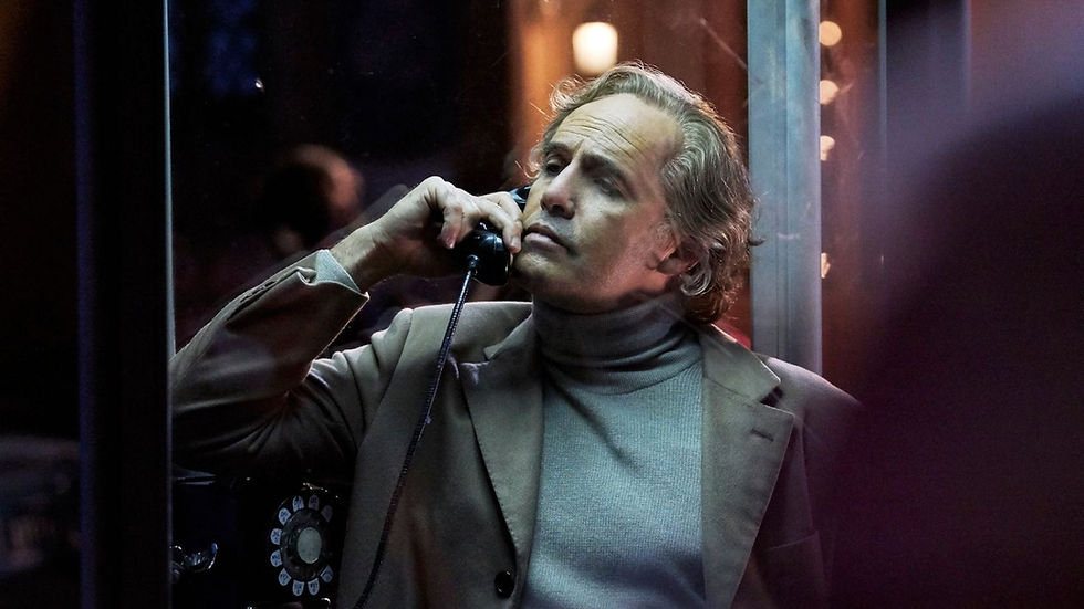 Man in a beige suit uses a rotary phone in a dimly-lit booth, looking contemplative. Warm lighting and blurred background.