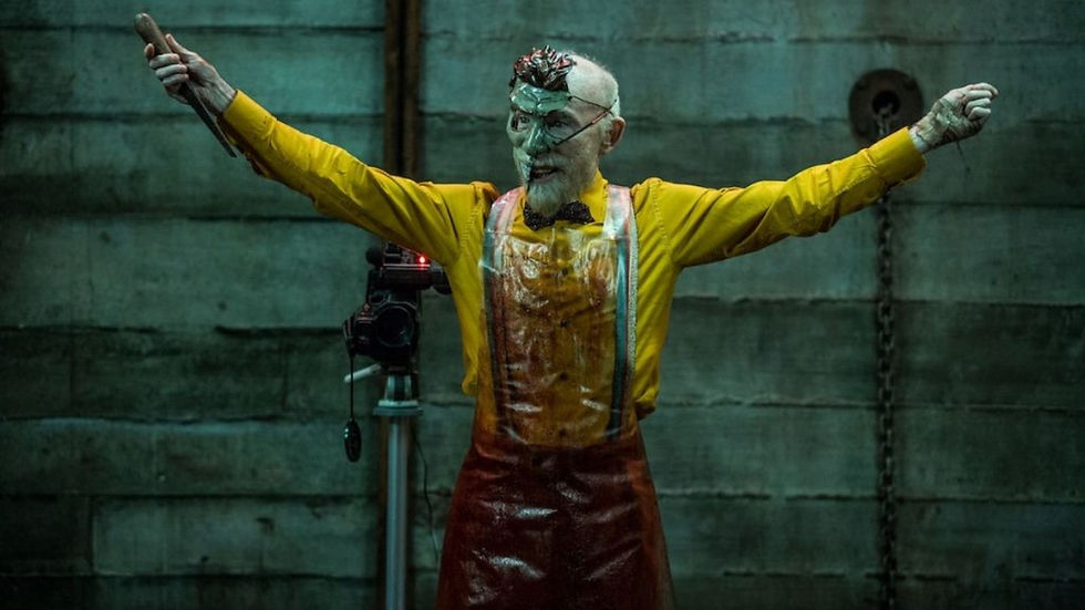 Man in yellow shirt and red apron with arms outstretched, wearing a mask. Industrial setting with camera, evokes eerie mood.