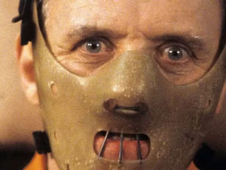 Close-up of a person wearing a beige restraint mask with metal bars, eyes wide open. The background is blurred, creating a tense atmosphere.
