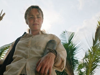 Woman in beige shirt holds a spear, standing against a backdrop of palm trees and blue sky, conveying a determined mood.
