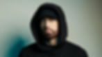 Man in black hoodie and cap, looking serious against a gradient backdrop. Pale blue eyes, light beard. Mood is intense and focused.