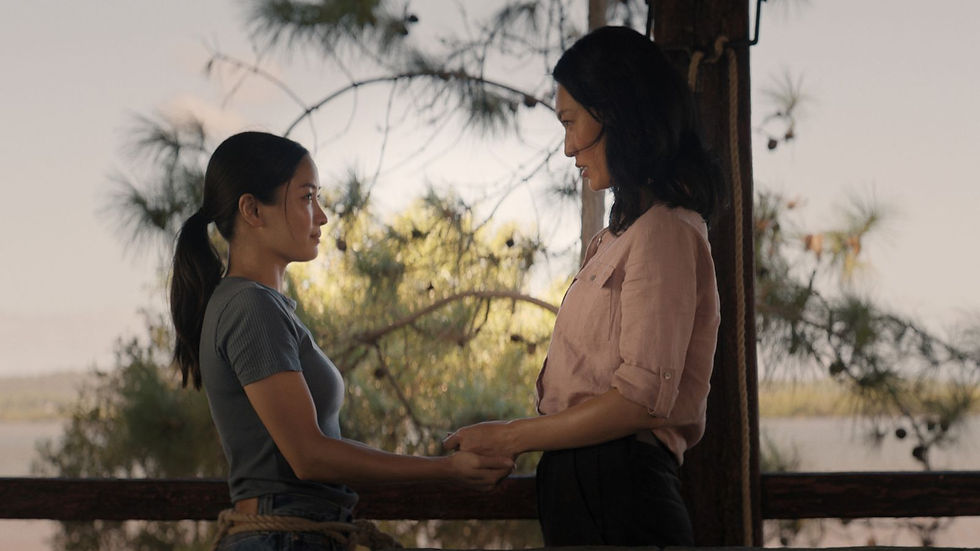 Two women stand facing each other holding hands on a porch. Trees and a lake are in the background, creating a serene atmosphere.