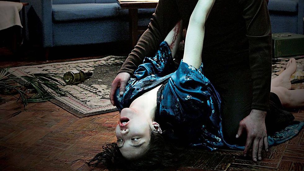 Man kneels over a pale woman lying upside down on a wooden floor in a dark room, creating a tense horror scene.