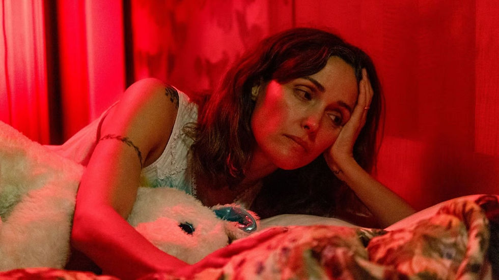 A woman with a tired expression lies on a bed, leaning on her hand. The room is bathed in red light, with a floral-patterned blanket.