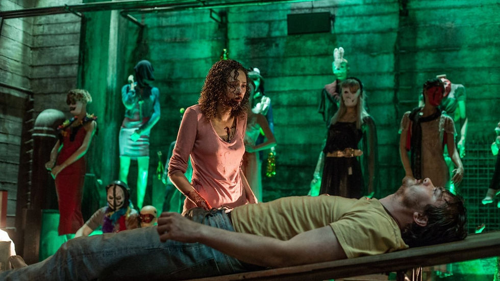 Woman in a pink top, looking distressed, stands over a man on a table. Green-lit room with masked mannequins in the background.