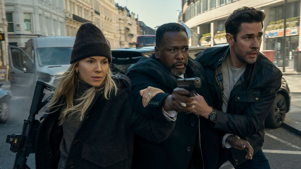 Three tense people move through a city street, one aiming a handgun, with a rifle on the woman’s back and cars behind.
