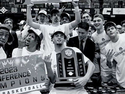 Basketball team celebrates 2023 Conference Champions victory. Players in white shirts hold trophy and sign, displaying excitement and joy.