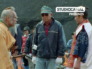 Three men stand outdoors; two in traditional robes facing a man in a hat and jacket. People watch in the background. Mood is tense.