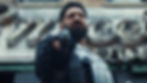 Bearded man in tactical gear points a gun towards the camera, standing in front of a vintage storefront sign. Intense expression, urban setting.