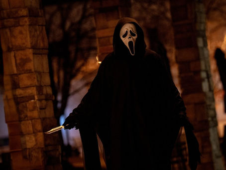 Person in a black cloak and white mask holds a knife in a dimly lit urban setting, creating a suspenseful mood. Brick and stone pillars visible.