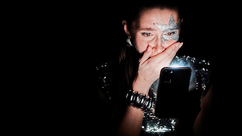 Woman with silver star face paint covers her mouth while looking at a glowing phone in a dark, dramatic setting.