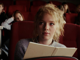 A young woman with blonde hair, holding a book, sits in a row of red theater seats. Others in the background appear relaxed or bored.