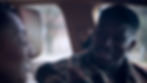 Two men talking in a car, one smiling in a patterned jacket, with beige seats and soft daylight outside.