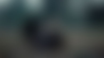 Man in brown jacket sits on fallen leaves in a graveyard, appearing contemplative. Tombstones and bare trees fill the misty background.