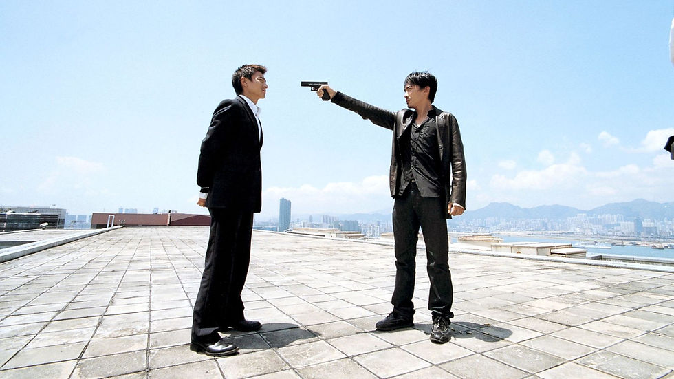 Man in black points a handgun at a suited man on a rooftop, with bright blue sky and city skyline behind them.