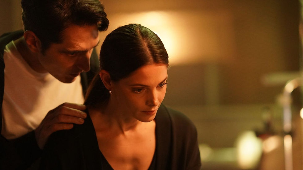 A man and woman in a dimly lit room, the man rests his hand on her shoulder. Both appear focused and serious, with warm light in the background.