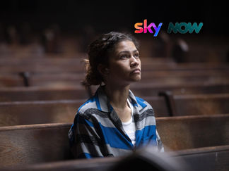 A person in a blue striped shirt sits alone on a wooden bench in a dimly lit space, looking contemplative and thoughtful.