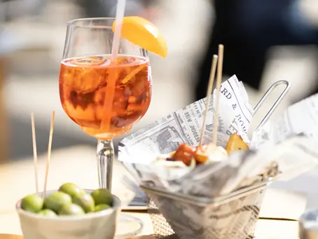 A sunlit close-up of an Aperol Spritz in a stemmed glass with ice, an orange slice, and a straw. Beside it sits a bowl of green olives. A basket of newspaper-wrapped snacks is blurred in the background.