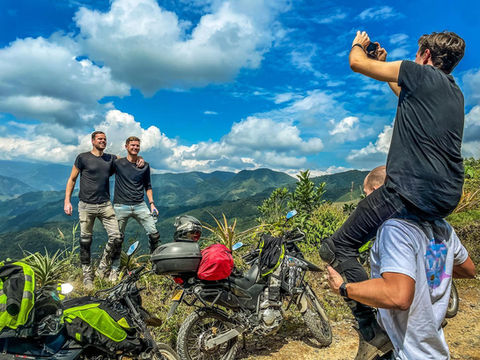 Three riders posing on a mountain viewpoint with motorcycles in the background.