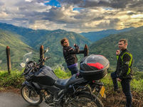 Two riders posing beside motorcycles with dramatic sunset sky over Colombian Andes.