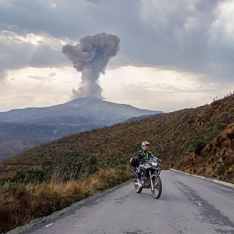 Motorcyclist riding with Nevado del Ruiz volcano erupting in background, Colombian Andes.