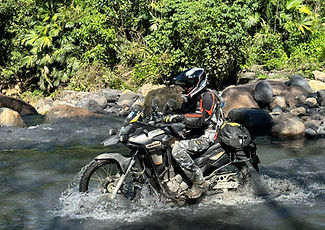 Dual-sport rider on a Honda XRE300 riding through a rocky stream surrounded by dense tropical forest in Colombia.