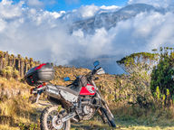 Dual-sport motorcycle parked at a high-altitude viewpoint with clouds and mountains.