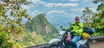 Smiling rider with Suzuki DR650 at a high Andean lookout with dramatic peaks behind.