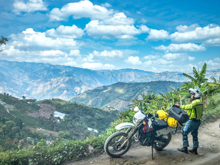 Rider and Suzuki DR650 at a Coffee Region overlook with blue skies and layered Andean valleys.